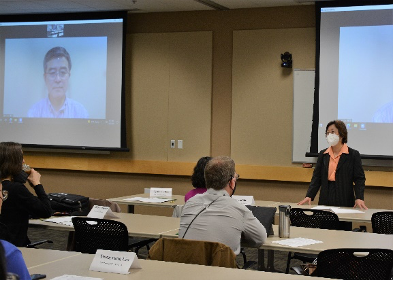 people sitting in a room at a conference; one person lecturing, wearing a mask