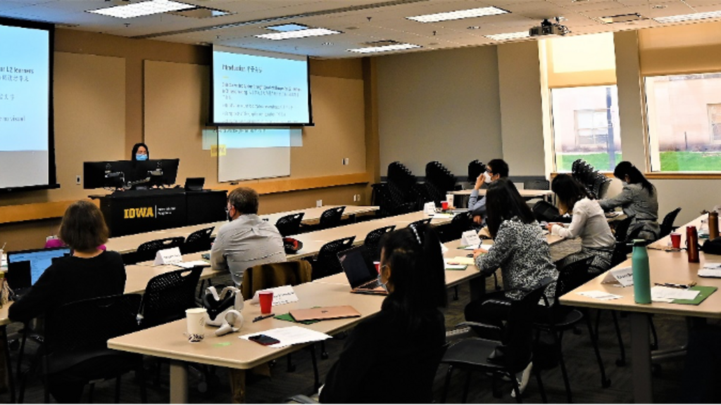 people sitting in a room at a conference; one person lecturing, wearing a mask